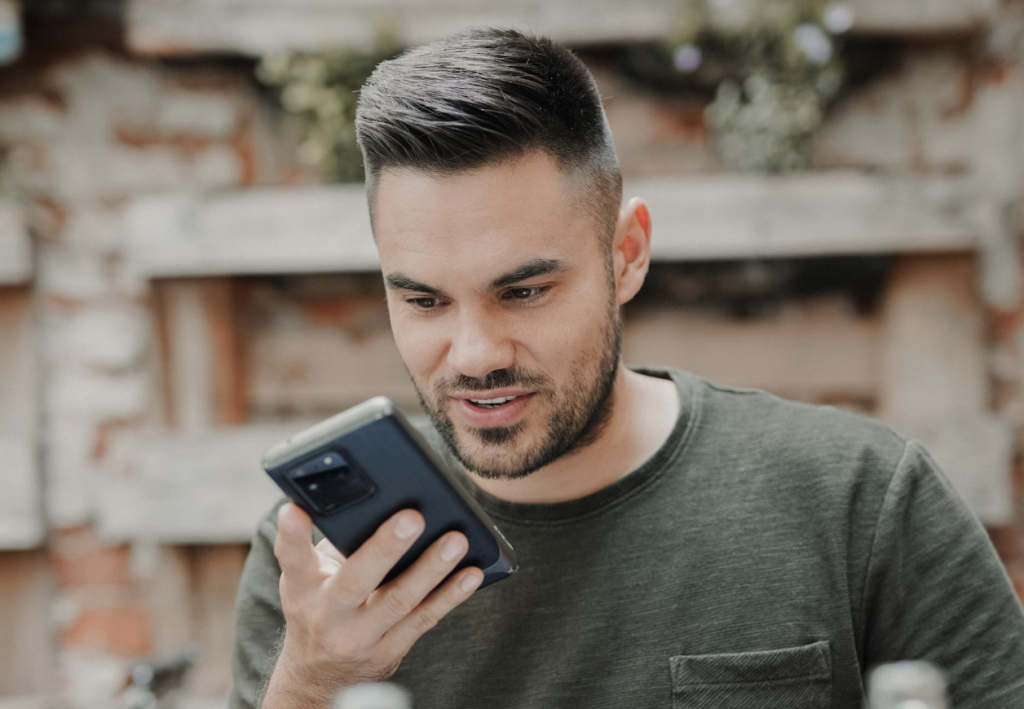 Man talking into the phone sending a voice message, sitting in a caffee work and travelling with a smartwatch, two drinks and leisure cloth. He is communicating via his smartphone.